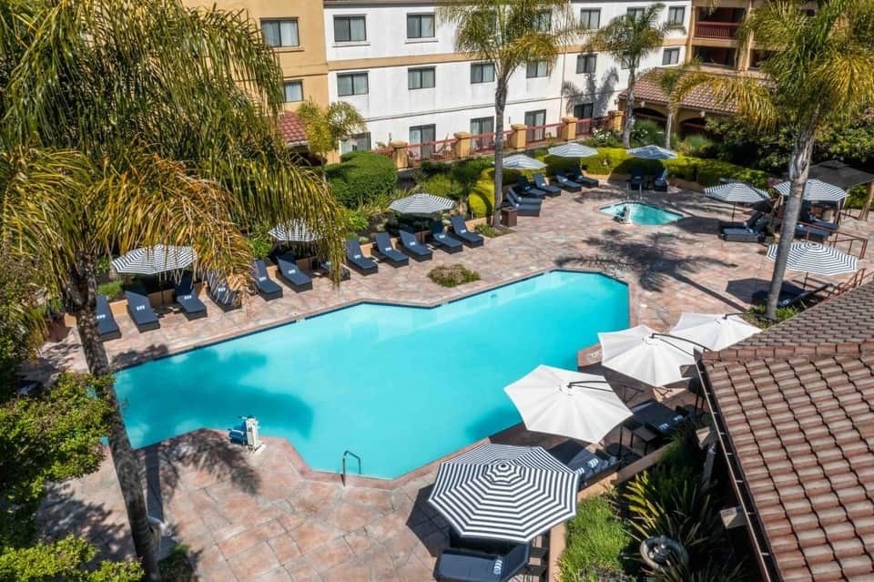 Resort pool complex with turquoise water, lounge chairs, striped umbrellas, palm trees, and surrounding residential buildings