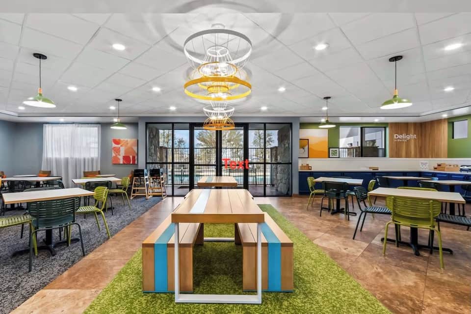 Bright communal workspace with long wooden tables, colorful chairs, large windows, and hanging pendant lights overlooking balcony