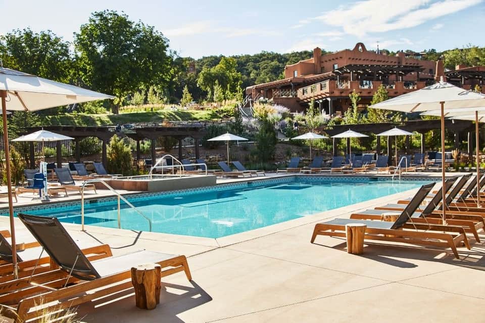 Resort pool deck with blue water, wooden loungers, white umbrellas, and rustic buildings