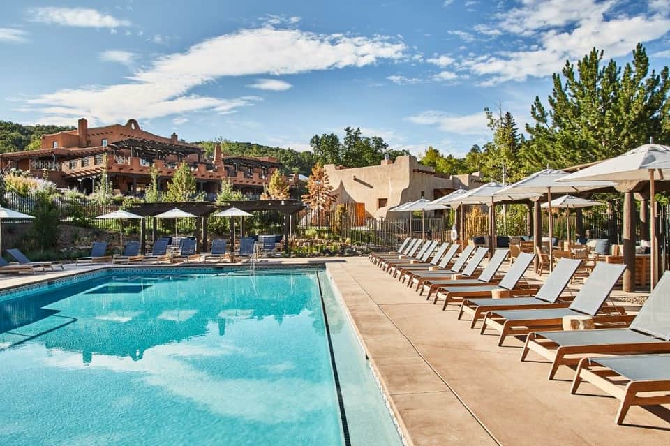 Resort swimming pool with wooden loungers, white umbrellas, and terracotta buildings in background