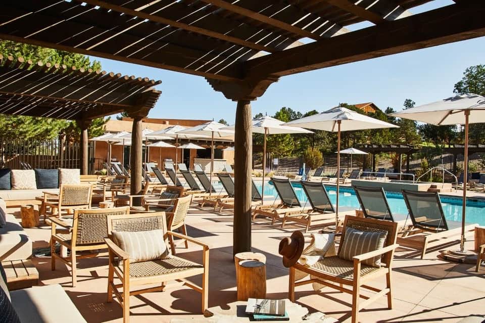 Luxury poolside lounge area with wooden pergolas, cushioned chairs, umbrellas, and resort buildings under clear blue sky