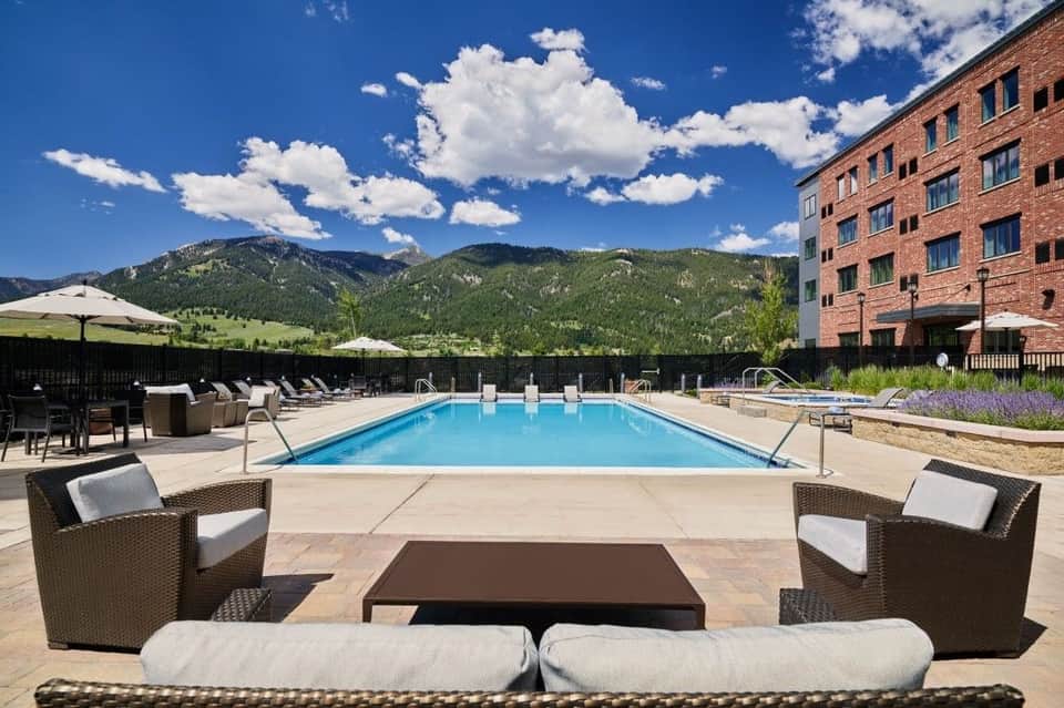Resort pool with mountain views, surrounded by lounge chairs, umbrellas, and a brick building with purple flowers in landscaping