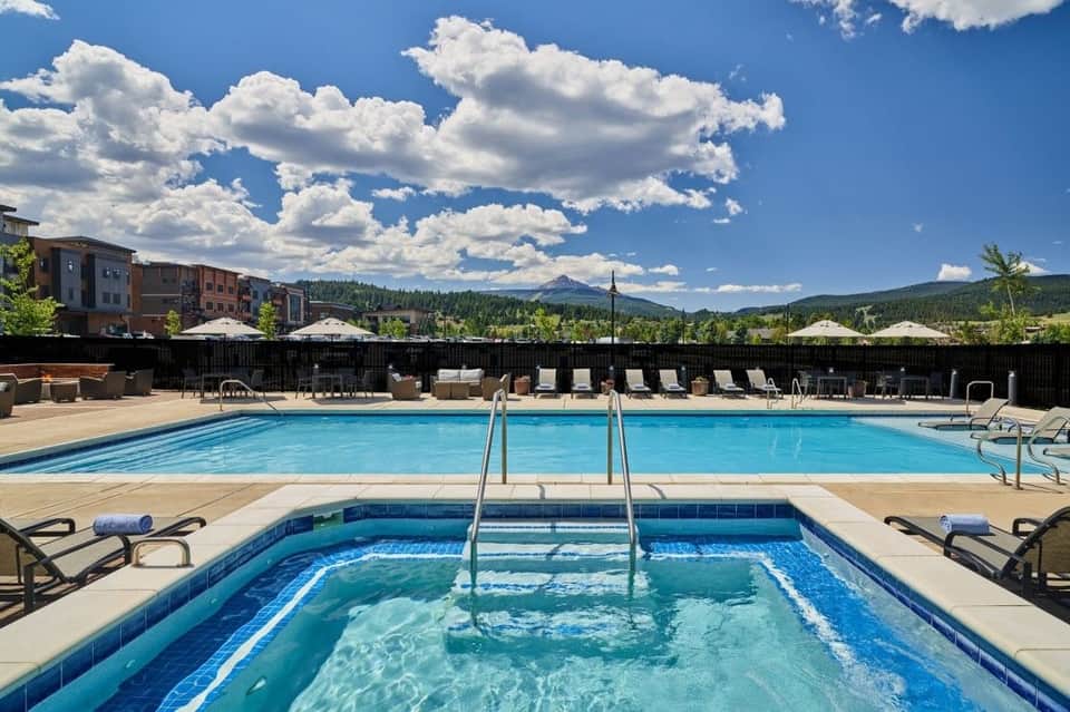 Resort pool area with mountain views, lounge chairs, umbrellas, and spa pool under blue sky