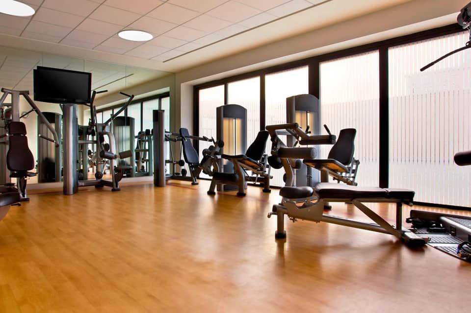 Well-lit fitness center with cardio and strength training equipment, wooden flooring, and large windows