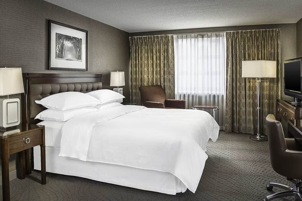 Upscale hotel suite with king bed, sitting area, elegant curtains, and taupe textured wallpaper throughout
