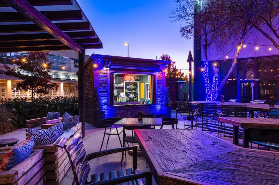 Evening courtyard with purple ambient lighting, wooden seating, bar counter, and decorated trees