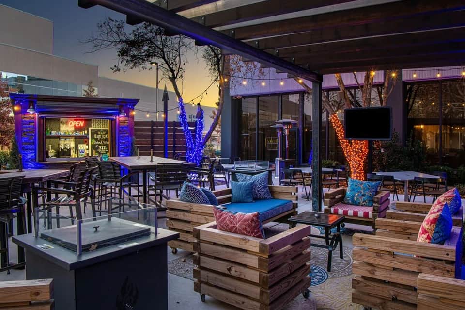 Modern rooftop lounge at dusk with wooden platforms, blue lighting, fire pit, and bar window