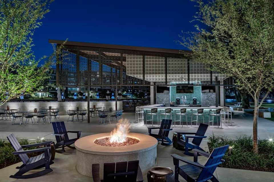 Nighttime outdoor lounge with fire pit, blue chairs, pergola, and bar area under starlit sky