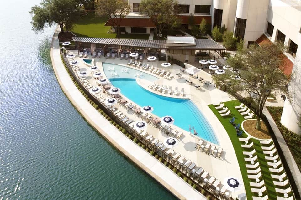 Aerial view of waterfront resort with curved pool, white umbrellas, lounge chairs, and manicured lawn