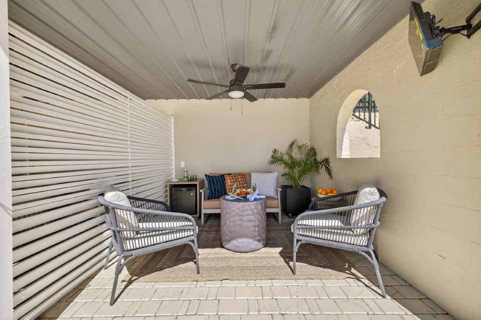 Covered patio lounge area with wicker chairs, potted plants, and modern furniture facing a stucco wall