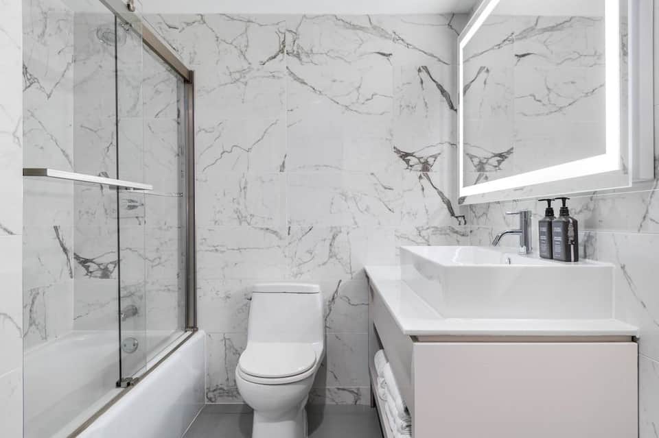 Modern bathroom with marble-tiled walls, glass-enclosed shower, white fixtures, and LED mirror