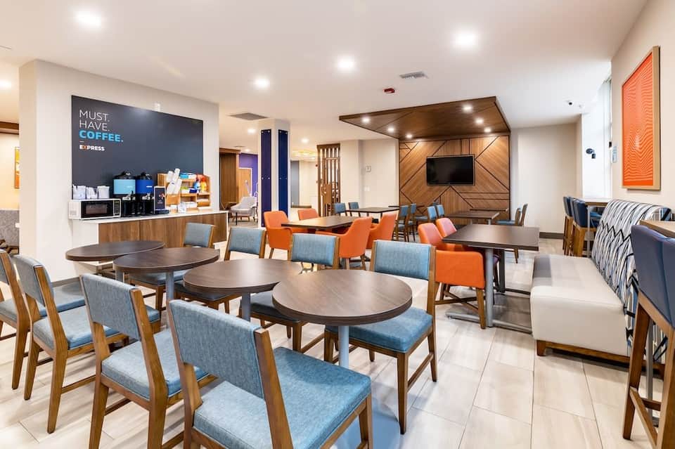 Modern hotel lobby and lounge area with round tables, blue and orange accent chairs, coffee counter, and wood-paneled entertainment wall