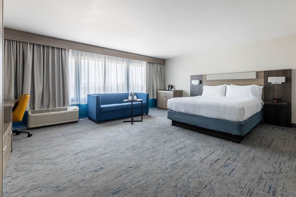 Spacious hotel suite with king bed, blue sofa seating area, floor-to-ceiling windows, and modern wood accents