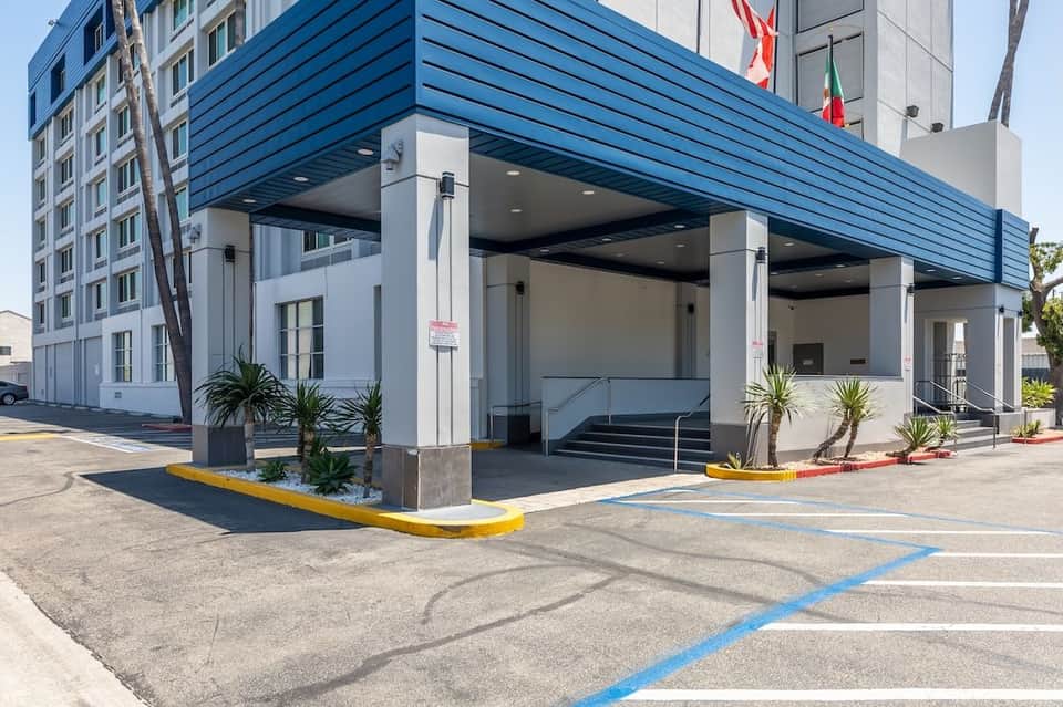 Modern hotel entrance with blue metal canopy, white columns, glass doors, and tropical landscaping