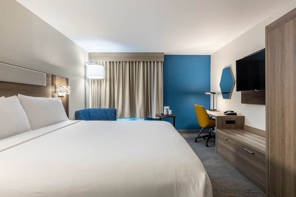 Modern hotel bedroom with blue accent wall, white bed, desk with yellow chair, and wall-mounted TV