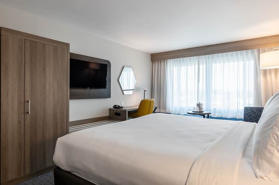 Spacious hotel suite with two beds, wood wardrobe, desk area, yellow chair, and sheer curtains