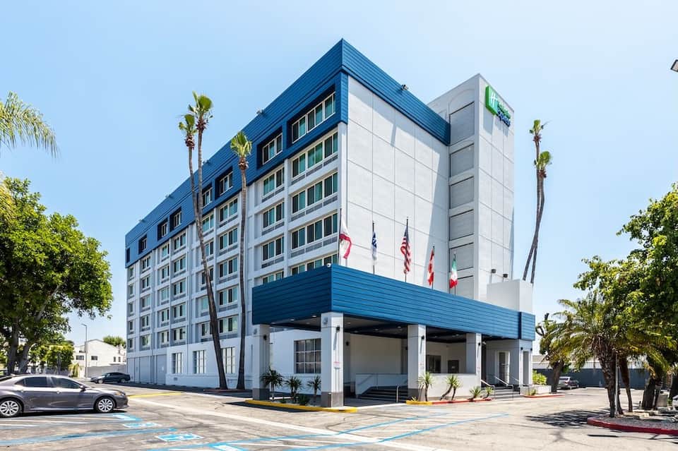 Modern six-story hotel building exterior with blue and white facade, palm trees, and ground-level entrance
