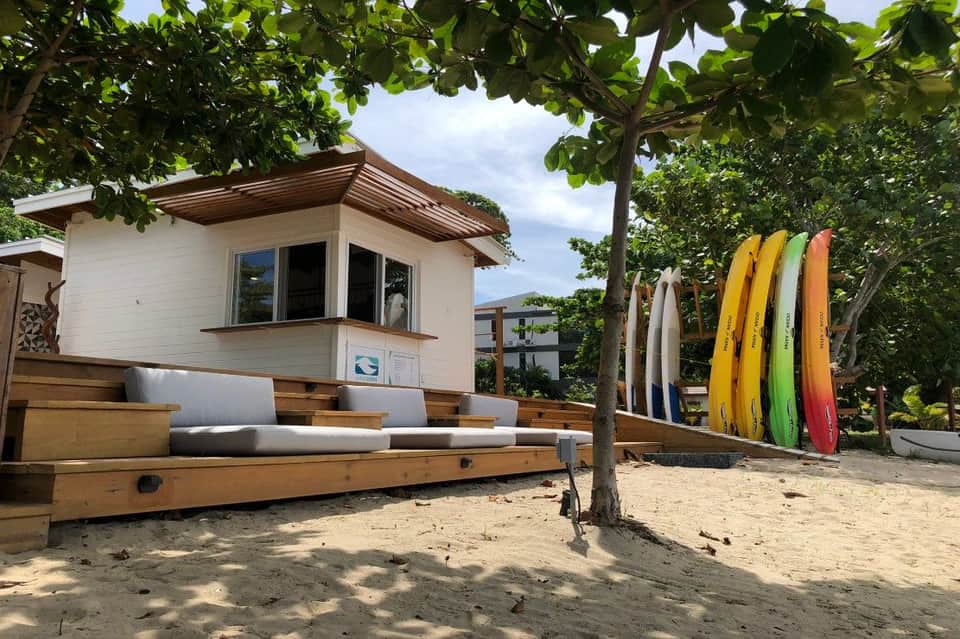 Beach shack with white walls and colorful surfboards leaning against trees on sandy ground