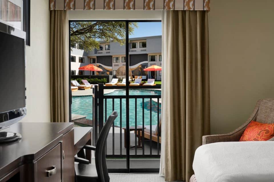 Hotel room with desk, bed, and balcony overlooking pool area with umbrellas and lounge chairs