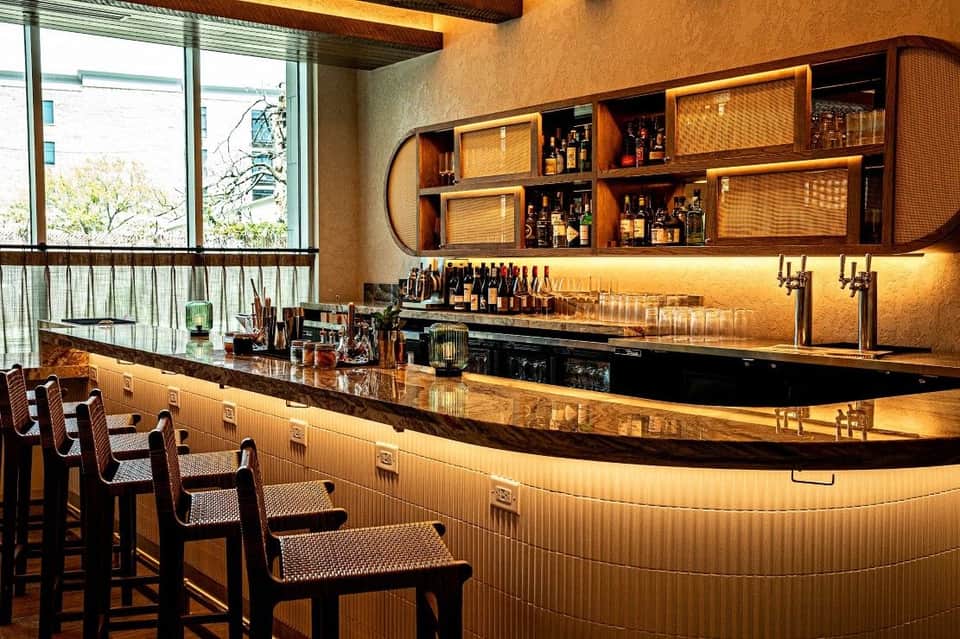 Upscale hotel bar with curved counter, wooden shelving, warm amber lighting, and bar seating overlooking city views