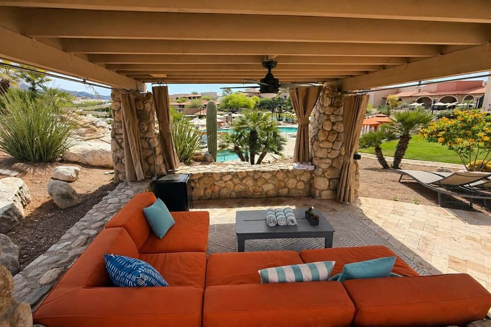Desert resort cabana with orange sectional seating, stone pillars, tan curtains, and pool view in background