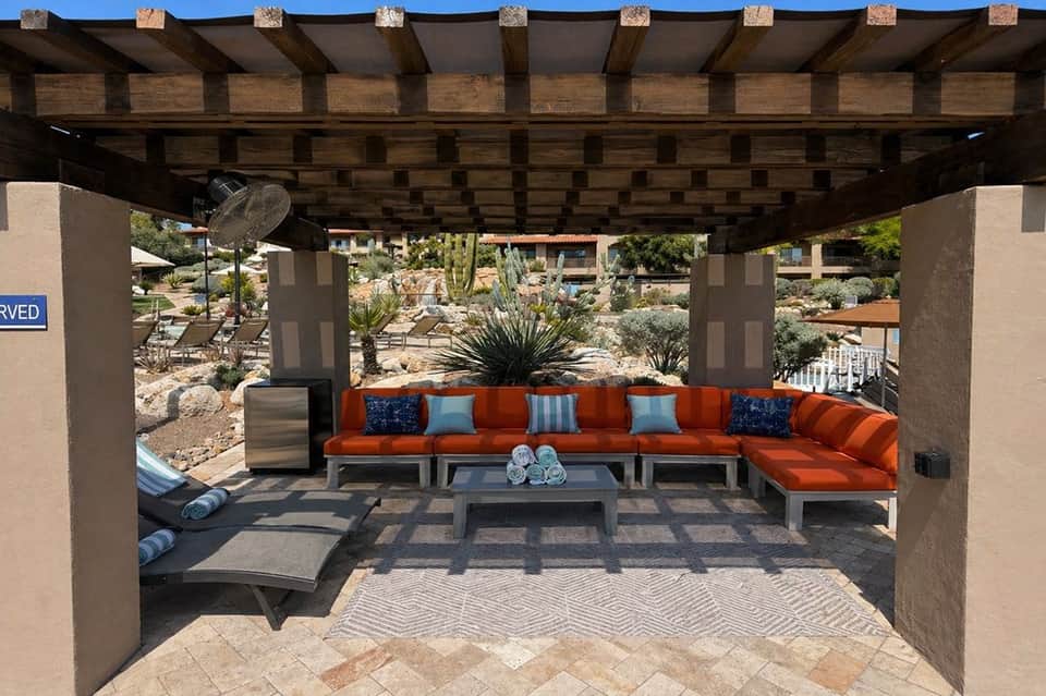 Reserved cabana with orange sectional sofa, blue pillows, and gray coffee table under wooden pergola