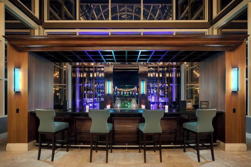 Modern hotel bar with purple ambient lighting, wooden paneling, and four upholstered bar stools facing illuminated shelving