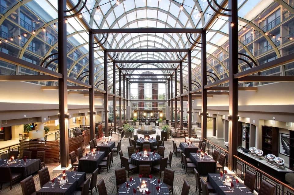 Grand covered dining hall with industrial glass ceiling, black tables set for events, and architectural steel framework