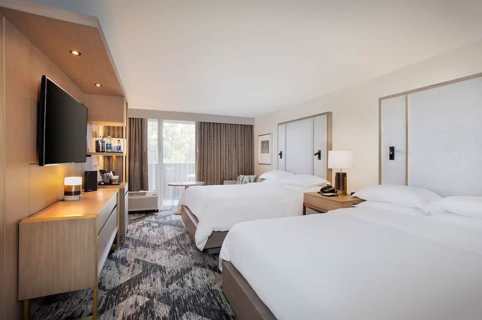 Contemporary hotel room with two queen beds, wood accents, flat-screen TV, and floor-to-ceiling windows with sheer curtains