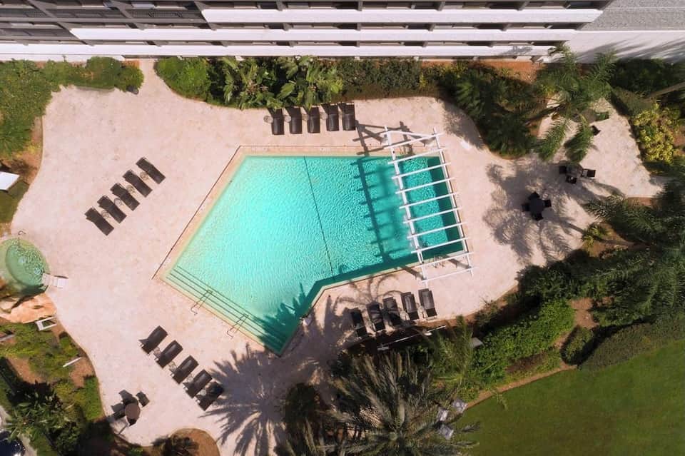 Aerial view of resort pool with turquoise water, lounge chairs, sandy deck, and surrounding tropical landscaping