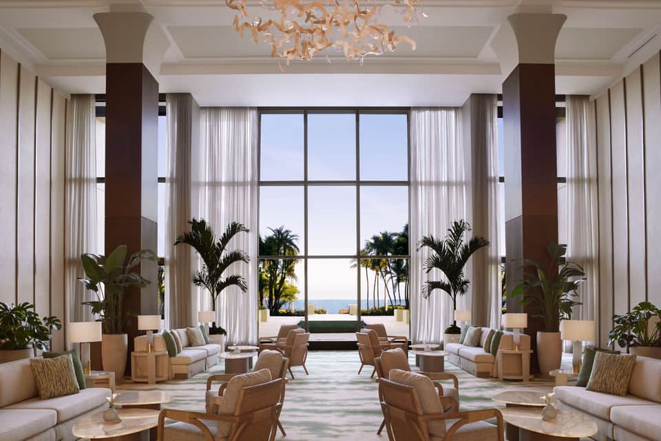 Luxurious lobby with tall windows, palm plants, cream furnishings, and ocean views