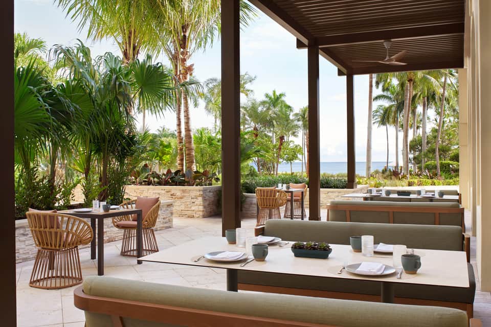 Covered oceanfront dining area with wooden chairs, ocean views, and lush palm landscaping