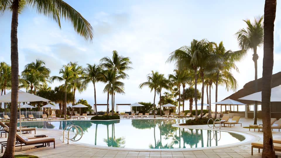 Large lagoon-style pool surrounded by white umbrellas, palm trees, and modern resort structures