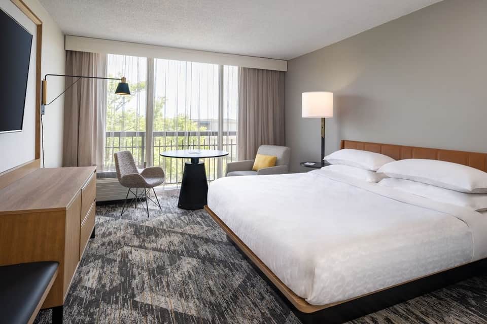 Hotel bedroom with king bed, wooden headboard, balcony access, work desk, and contemporary neutral décor