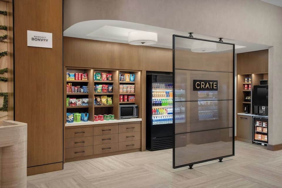 Modern hotel marketplace with wooden shelving, refrigerated beverage display, snacks, and glass partition labeled CRATE