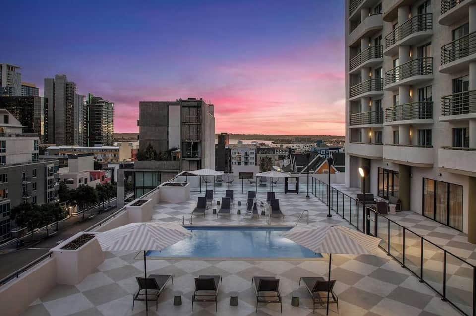 Rooftop pool at dusk with pink sunset sky, striped umbrellas, and city skyline backdrop