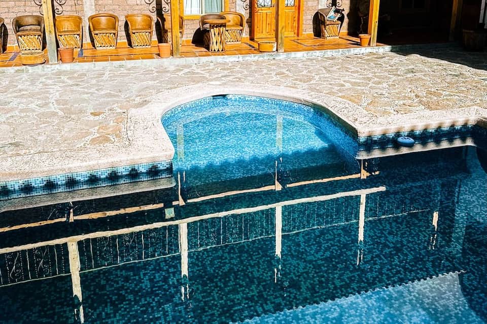 Kidney-shaped pool with blue mosaic tiles, stone decking, and wooden chairs beside building