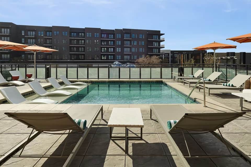 Rooftop pool deck with loungers, orange umbrellas, and modern dark apartment buildings in background