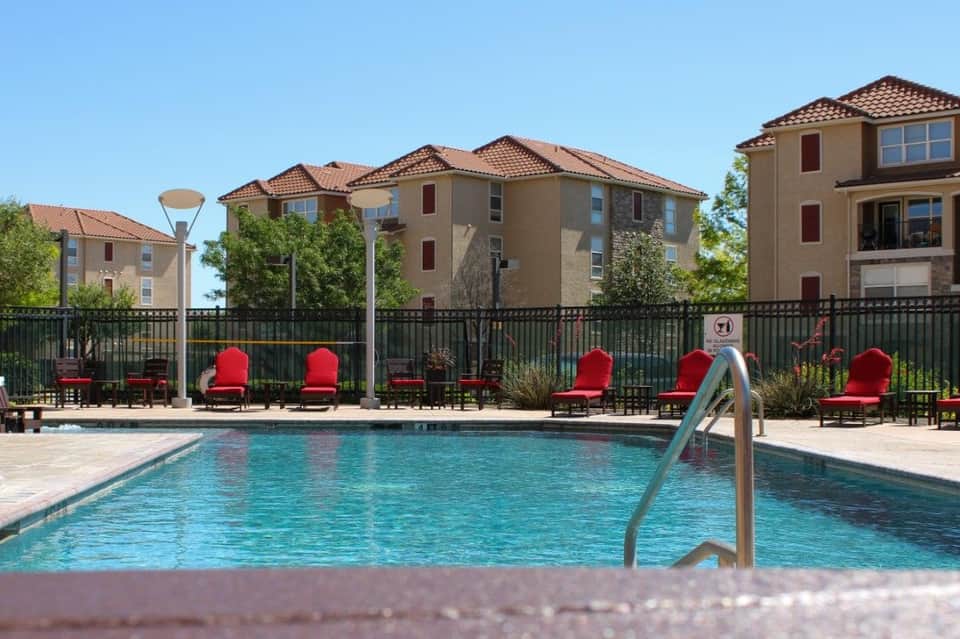 Swimming pool fronted by red cushioned lounge chairs and black fence with tan residential buildings behind