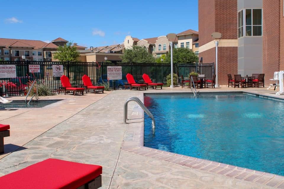Outdoor swimming pool with blue water, red lounge chairs, brick residential building, and black perimeter fence