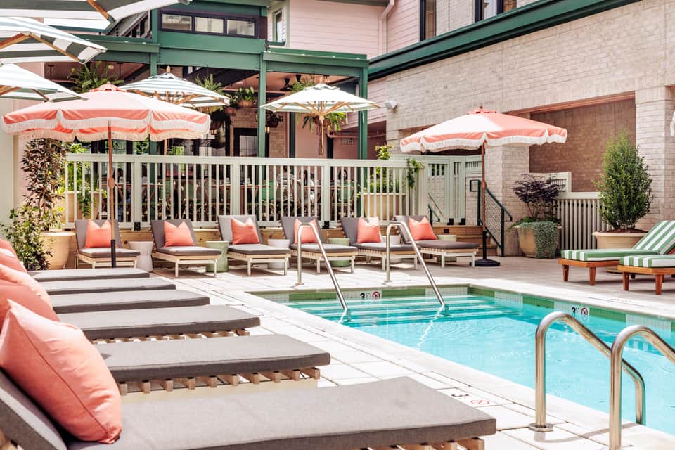 Resort pool deck with turquoise pool, pink umbrellas, lounge chairs with coral cushions, and white railing
