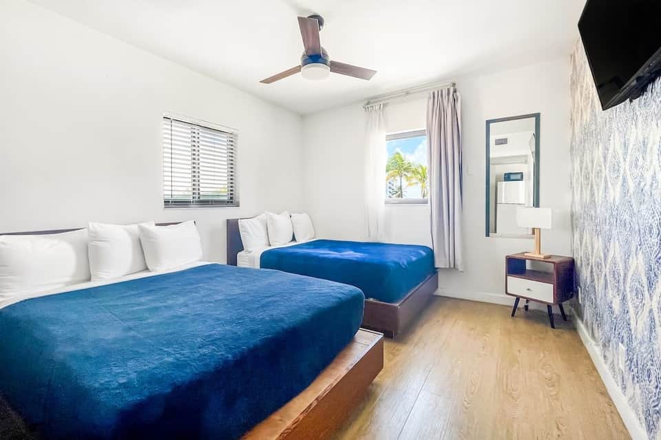 Hotel bedroom with two blue queen beds, ceiling fan, white walls, and palm tree view through window