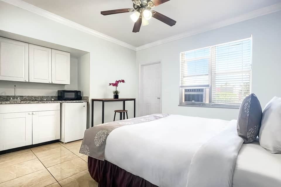 Modern hotel room with queen bed, kitchenette, ceiling fan, and large window with shutters