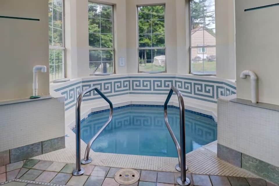 Indoor hot tub with Greek key tile pattern, metal handrails, large windows overlooking green landscape