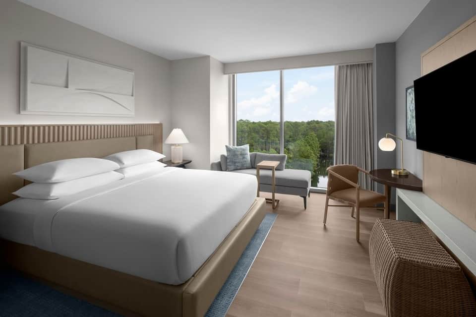 Modern hotel bedroom with white bedding, nature view windows, wood accent headboard, and contemporary furnishings