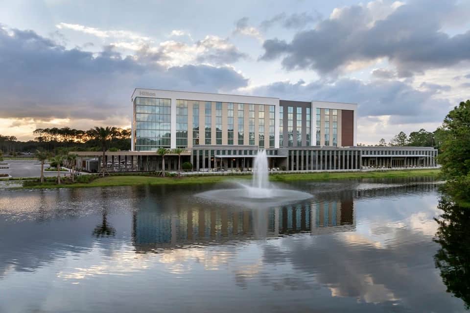 Contemporary Hilton hotel exterior reflecting in calm water with fountain, surrounded by green landscape at dusk