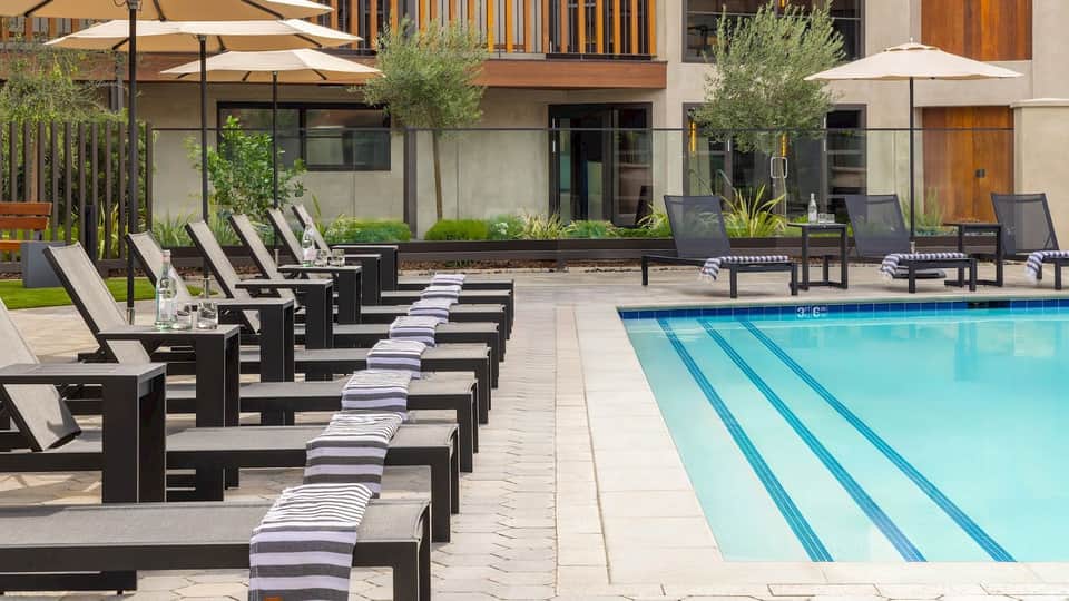 Resort pool deck with black lounge chairs, striped cushions, white umbrellas, and modern building architecture
