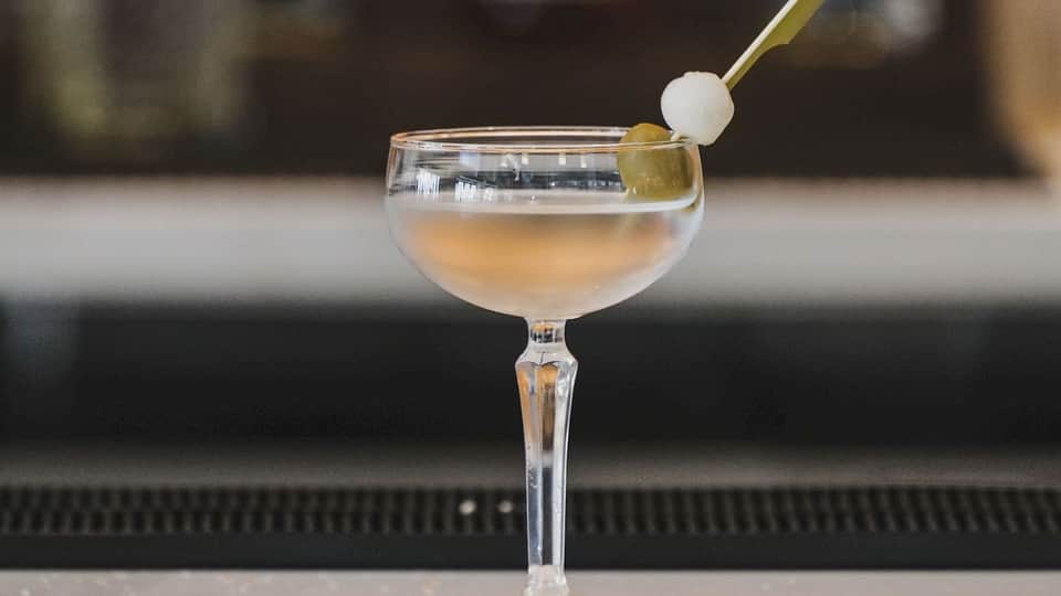 Craft cocktail in coupe glass with olive garnish and pearl onion on dark bar counter