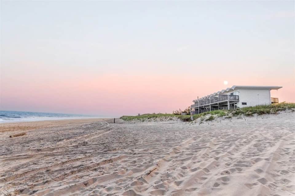 Beachfront house at dusk with pink sky, moon visible, sandy beach in foreground
