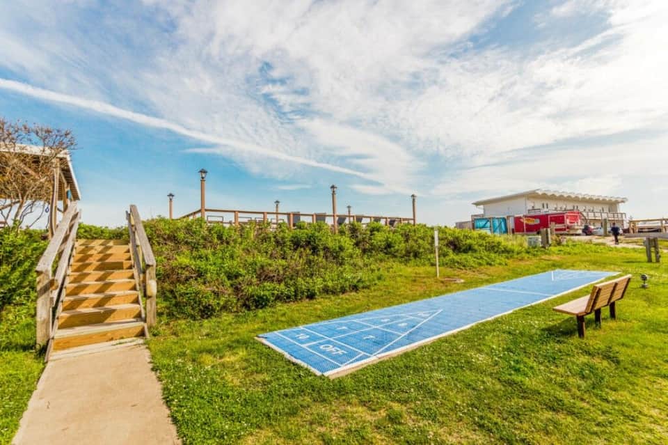 Grassy area with shuffleboard court, wooden walkway, and beach pavilion structures